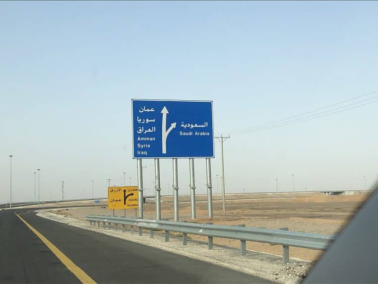 Route goudronnée à deux voies traversant un désert rocailleux, bordée de collines ocre sous un large ciel clair légèrement voilé.
