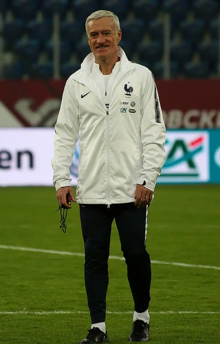 Didier Deschamps sourit en survêtement blanc de l’équipe de France sur la pelouse d’un stade, un sifflet à la main, lors d’un entraînement d’avant-match en 2018.