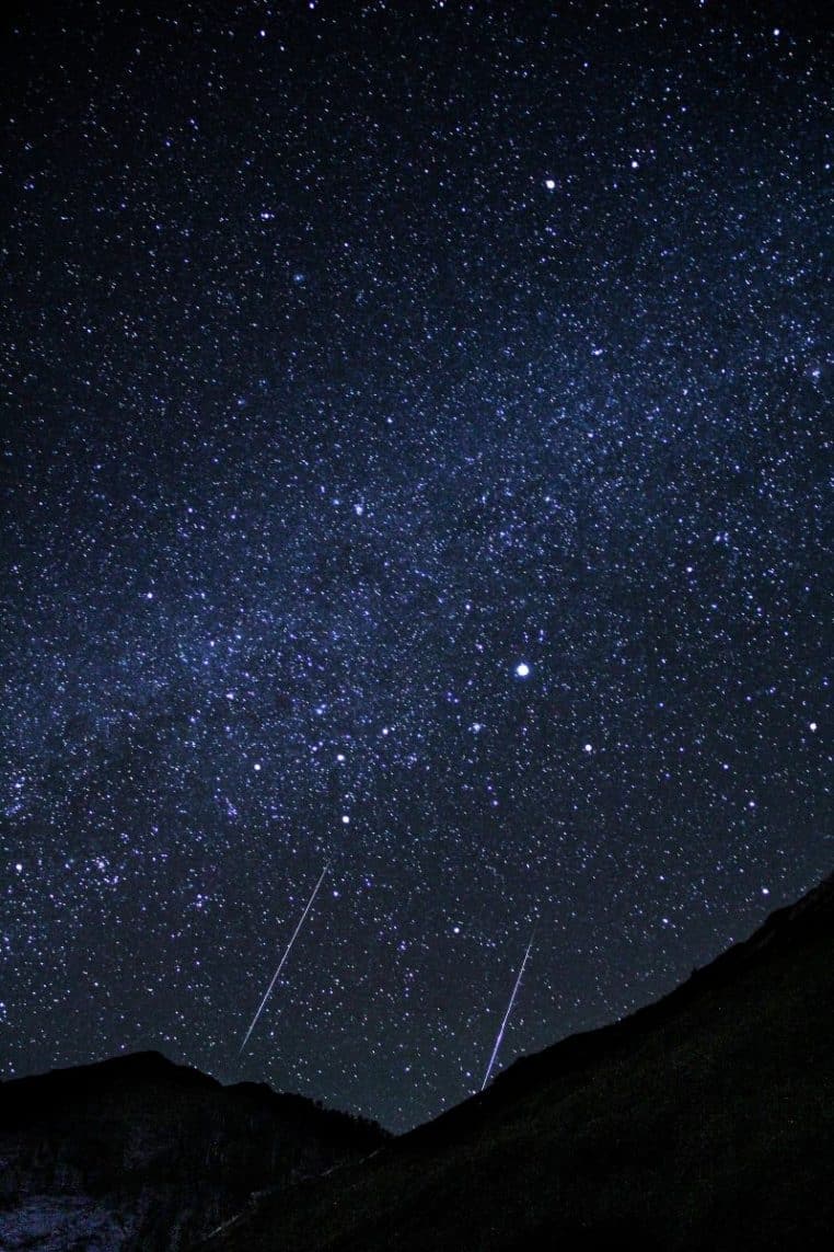Ciel de montagne très sombre rempli d’étoiles, traversé par deux longues traînées lumineuses parallèles de météores au-dessus des reliefs.