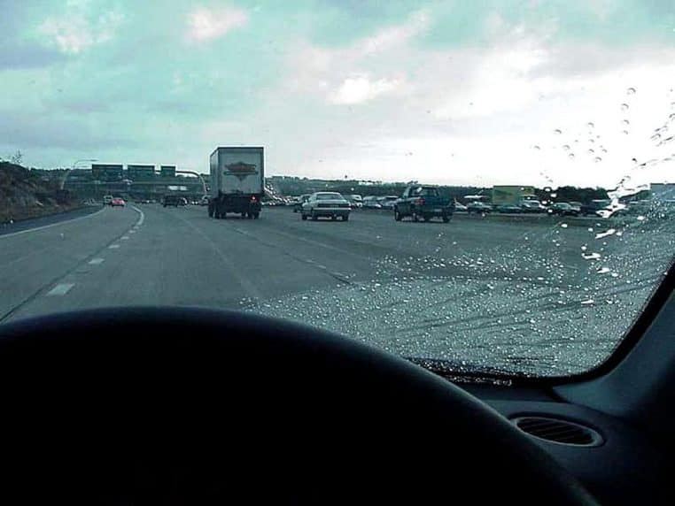 Route détrempée vue à travers un pare-brise mouillé, les voitures et les phares se devinent derrière des traînées de pluie sur la vitre.