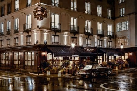 Façade nocturne du restaurant Drouant, au coin de la rue Gaillon, éclairages chauds reflétés sur la chaussée mouillée, terrasse végétalisée.