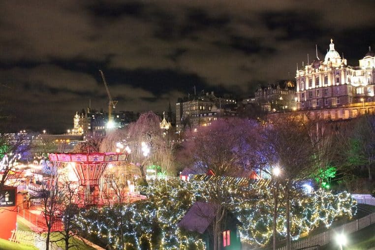Allée principale du marché de Noël, décorations lumineuses suspendues au-dessus des stands et foule se promenant entre les chalets.