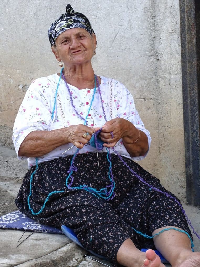 Femme âgée tricotant assise dehors dans une rue ensoleillée, pelote de laine et aiguilles visibles.