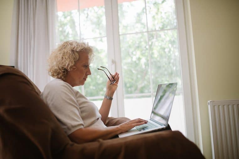 Femme âgée assise dans un fauteuil près d’une grande baie vitrée, tenant ses lunettes à la main tout en travaillant sur un ordinateur portable posé sur ses genoux.