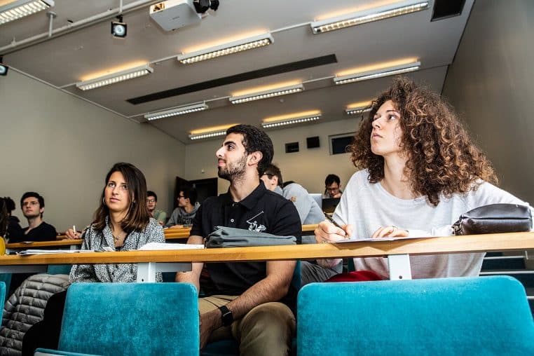 Salle de cours moderne à l’École polytechnique : des étudiants suivent un cours de mathématiques, rangées de tables et écran projeté visibles.