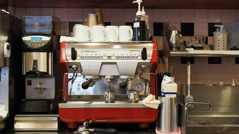 Machine expresso professionnelle rouge avec broyeur et tasses empilées sur le dessus, installée derrière un comptoir de café.