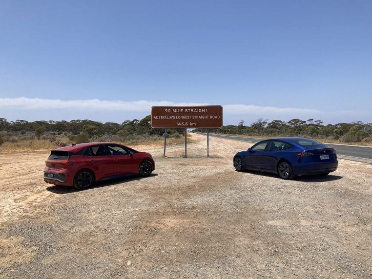 Panneau routier « 90 Mile Straight » encadré par deux voitures sur un bas-côté poussiéreux, avec la route qui s’enfonce dans le paysage.
