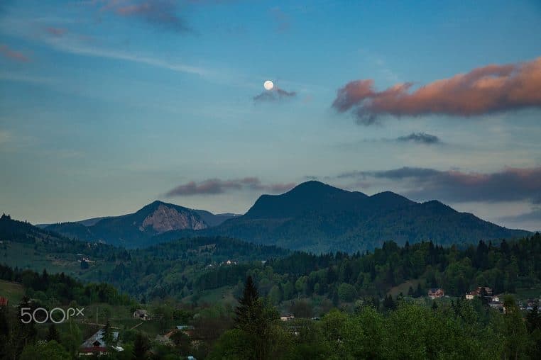 Pleine lune au-dessus d’un village de montagne, large plan paysager avec reliefs et ciel dégagé.