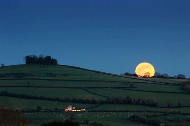 Pleine lune se levant au-dessus d’un relief anglais, vue large au crépuscule avec silhouettes et ciel clair.