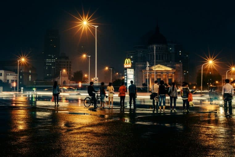 Grande avenue de nuit après la pluie, sol brillant de reflets, piétons alignés au feu rouge face aux phares et aux immeubles illuminés.