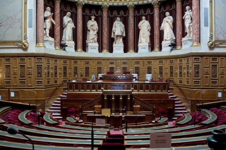 Vue large de l’hémicycle du Sénat français presque vide, montrant les bancs rouges et l’estrade centrale où se votent les lois sur les allocations et la fraude.