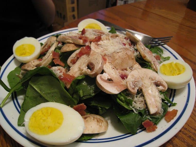 Salade d’épinards frais garnie d’œufs durs, de champignons et de petits morceaux de bacon, servie dans une assiette blanche.