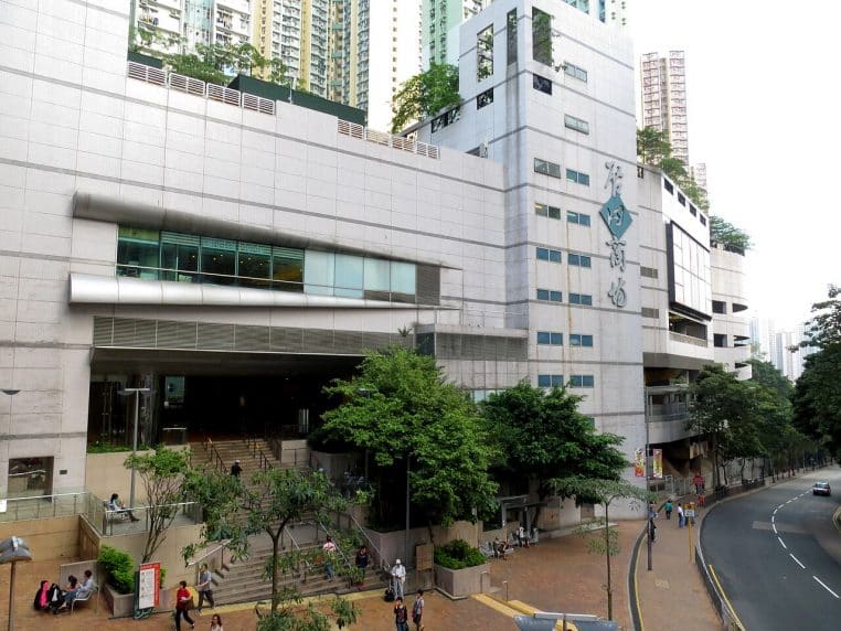 Grand centre commercial urbain longeant une route courbe, entrée vitrée en surplomb et piétons rassemblés près des escaliers.