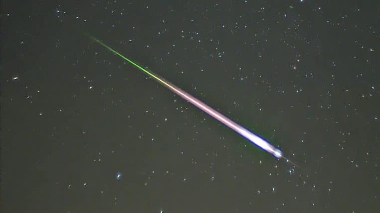 Gros plan d’une étoile filante des Léonides traversant obliquement un ciel nocturne étoilé, avec une traînée colorée nette allant du vert au rose.
