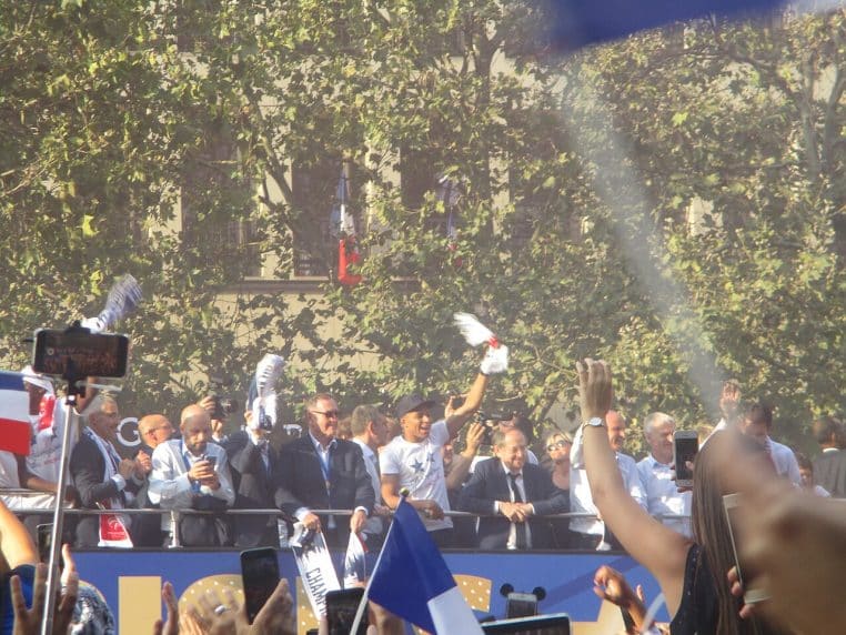 Les joueurs de l’équipe de France saluent une foule compacte depuis un bus à impériale sur les Champs-Élysées après le titre mondial 2018, avec Didier Deschamps et Kylian Mbappé visibles au premier plan.