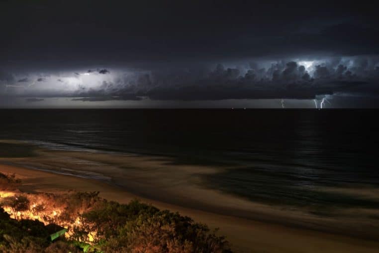 Orage nocturne avec éclairs au dessus de l’océan et nuages très sombres, vagues agitées et plage faiblement éclairée au premier plan.