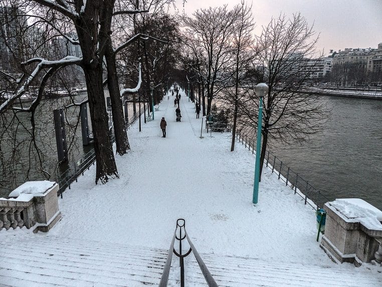 Promenade de l’île aux Cygnes recouverte de neige, quelques passants emmitouflés marchent entre les arbres, la Seine de part et d’autre renforce l’ambiance hivernale.