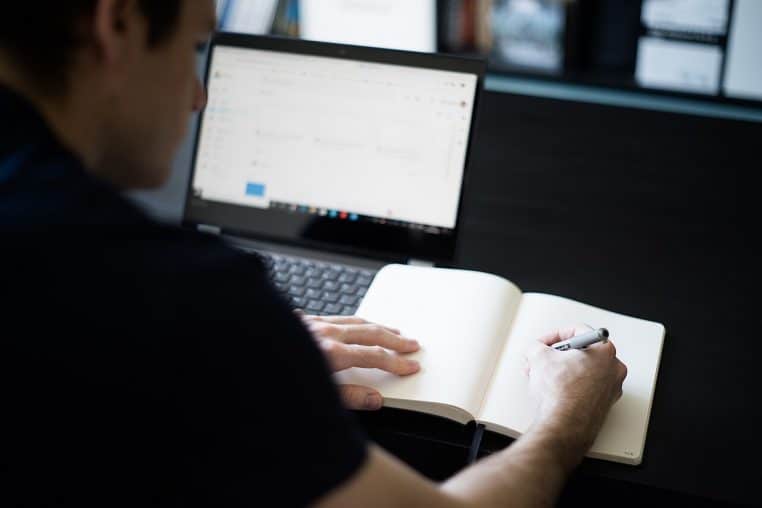 Homme prenant des notes devant un ordinateur portable posé sur une table basse, illustration d’un télétravail organisé depuis le domicile.
