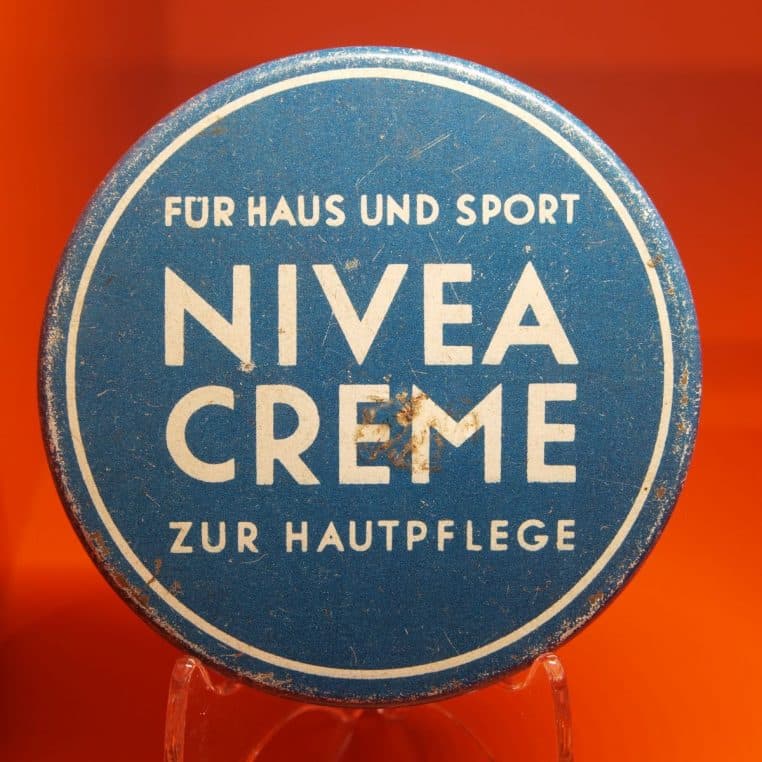 Gros plan sur une ancienne boîte ronde bleue de Nivea Creme exposée sur fond orange, illustrant l’héritage historique de la marque.