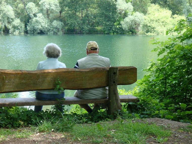 Deux personnes âgées assises sur un banc en bois face à un lac, vue de dos, illustrant une retraite paisible après une longue carrière professionnelle.