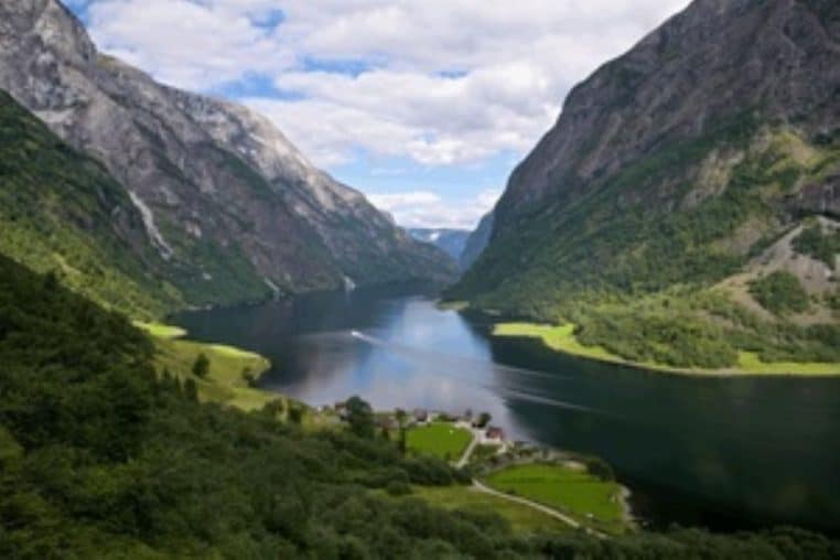 Large panorama d’un fjord norvégien avec un paquebot au loin, reliefs escarpés et eau lisse sous une lumière douce.