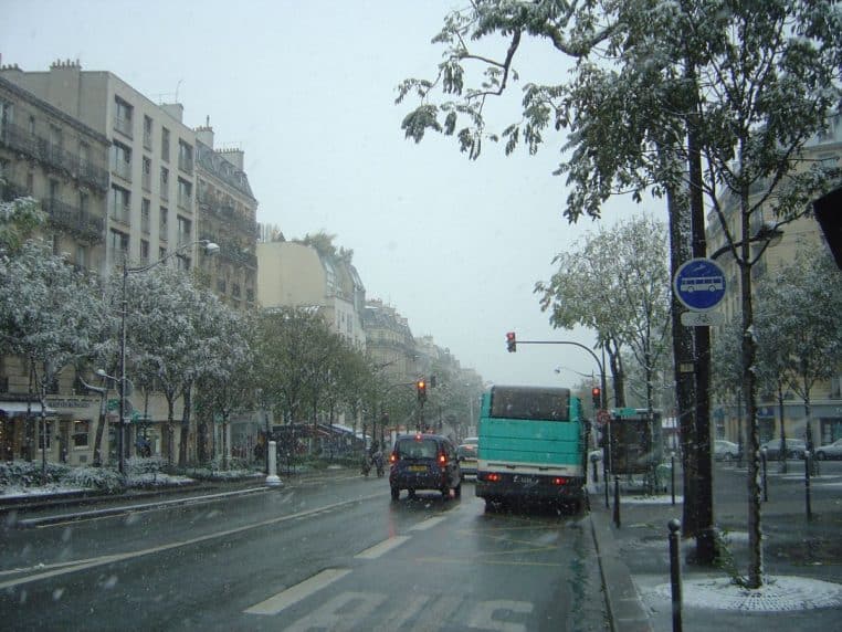 Avenue parisienne enneigée avec arbres nus, voitures stationnées et trottoir recouvert de neige, ambiance froide de début d’hiver.