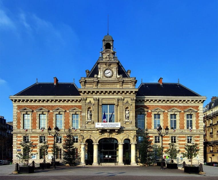 Façade monumentale de la mairie du 19ᵉ arrondissement de Paris, cadrée en plan large, représentant l’accès aux services d’état civil.