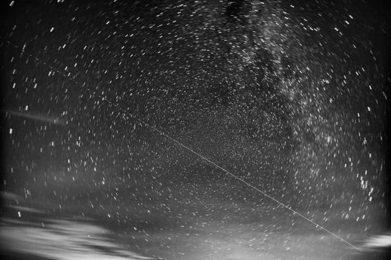Vue grand angle d’un ciel nocturne en noir et blanc avec traînée de météore et longues traces d’étoiles tournant autour du pôle céleste.