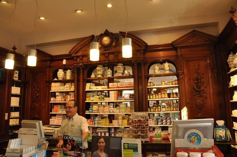 Intérieur lumineux d’une pharmacie parisienne avec comptoir, rayonnages et croix verte visible depuis la vitrine.
