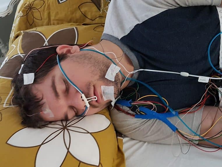Patient de profil équipé pour un enregistrement du sommeil, électrodes et câbles connectés à l’unité de mesure au chevet.