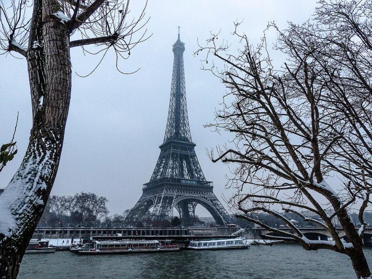 La tour Eiffel vue depuis le port de La Bourdonnais sous la neige, arbres dénudés, bateaux à quai et ciel uniforme donnant une impression de froid humide.