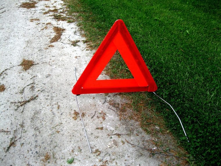Gros plan sur un triangle de présignalisation rouge posé à même le sol, entre un chemin clair et une bordure d’herbe verte bien contrastée