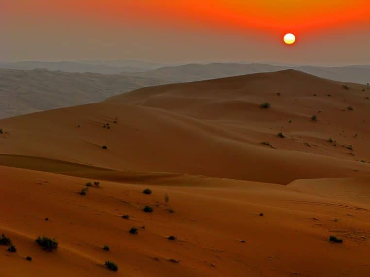 Paysage désertique au coucher du soleil avec de grandes dunes orangées et un disque solaire rouge brillant au-dessus de l’horizon.