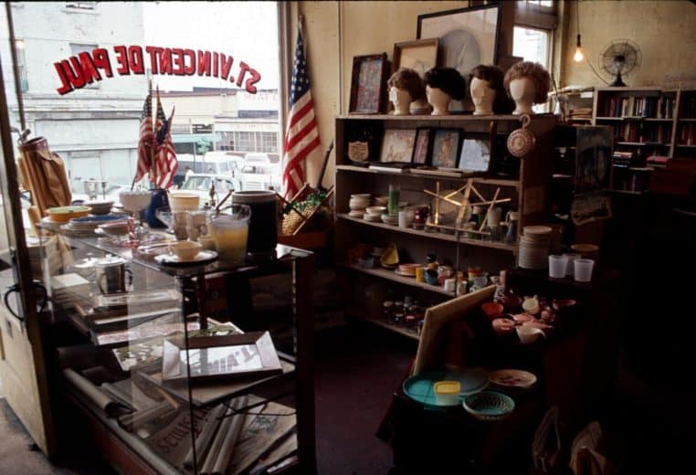 Vue d’ensemble d’une boutique Saint-Vincent-de-Paul en 1968, portants chargés de vêtements et ambiance rétro.