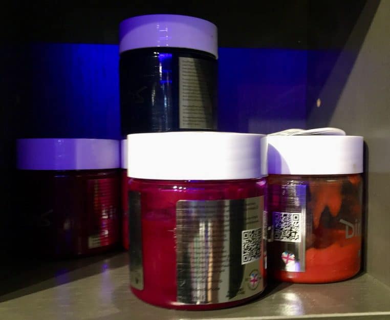 Pots de colorations capillaires semi-permanentes aux teintes vives rangés sur une étagère sombre éclairée par une lumière bleutée