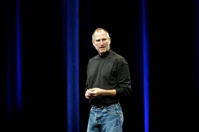Steve Jobs debout sur scène lors d’une keynote Apple, en col roulé noir, s’adressant au public devant un écran géant.