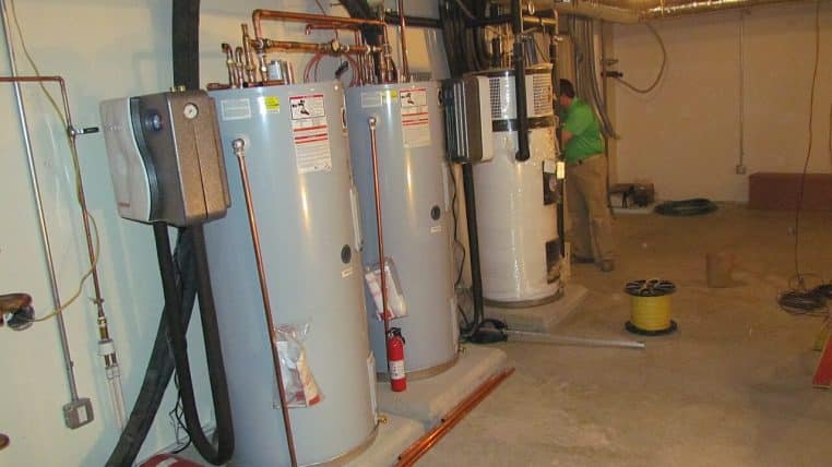 Inspecteur vérifiant un ballon d’eau chaude au sous-sol, avec tuyauteries apparentes sur un mur en pierre.