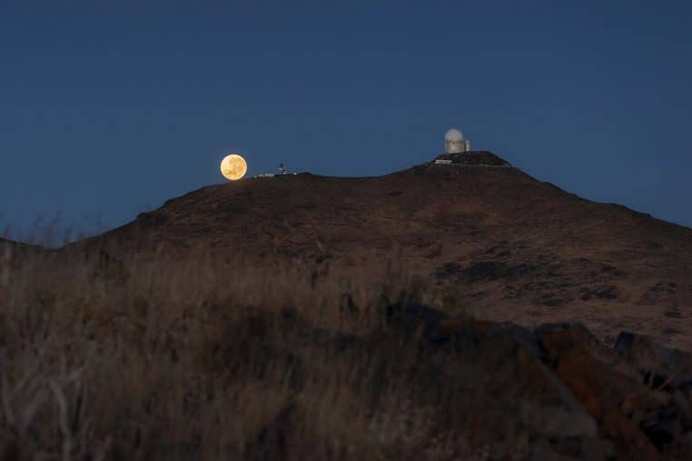 Super Lune posée sur une crête face aux coupoles de l’observatoire de La Silla au Chili, ciel clair et ton bleu nuit.