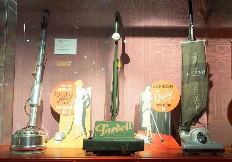 Aspirateur ancien exposé dans un musée de l’électricité, posé sur un sol clair devant un mur neutre.
