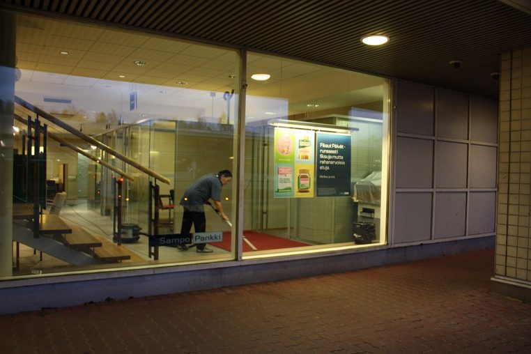 Jeune adulte passant l’aspirateur dans une chambre, manche incliné vers le sol, lit et mobilier visibles en arrière-plan.