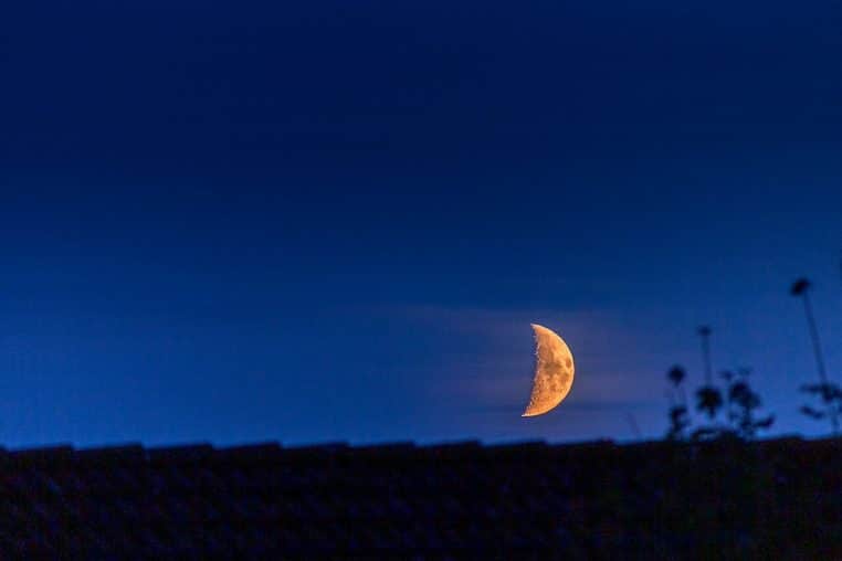 Fin croissant de Lune descendant derrière un toit suédois sous un voile de nuages, ciel indigo et ambiance calme en début de nuit.