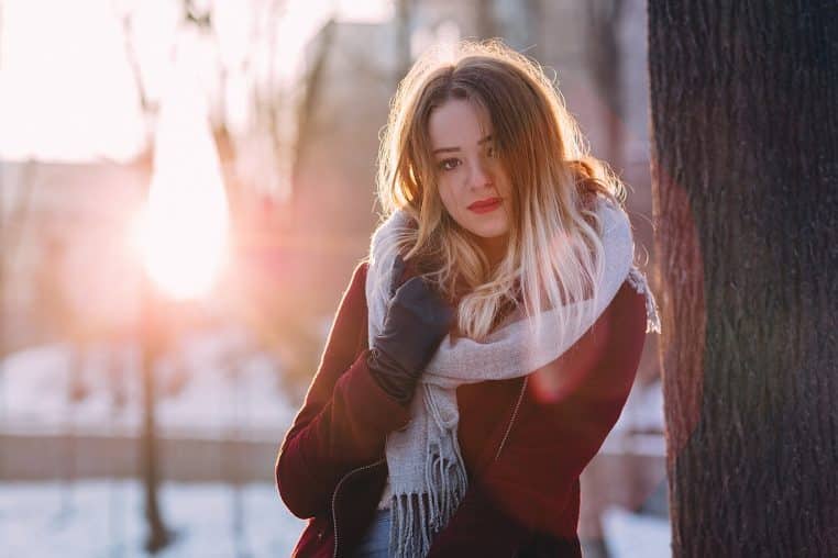 Jeune femme en manteau bordeaux et grande écharpe claire posant dehors dans la neige au coucher du soleil, ambiance hivernale chaleureuse.