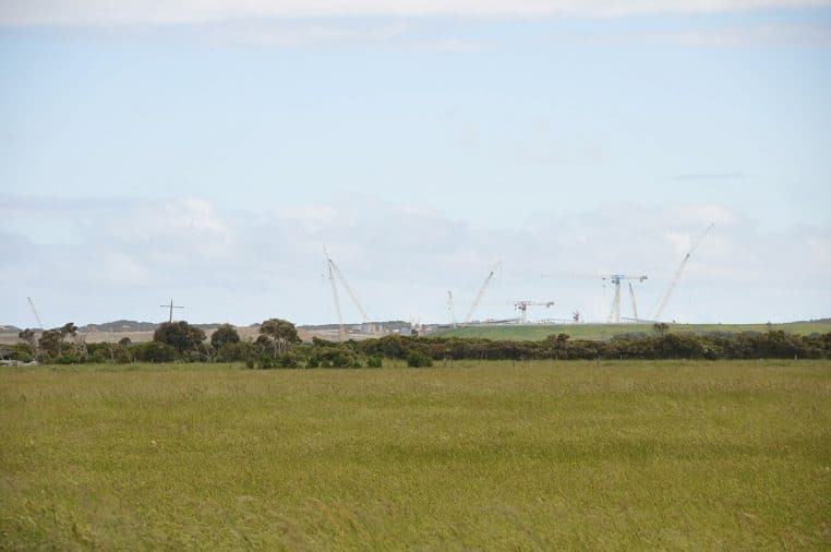 Chantier d’une usine de dessalement vu depuis une prairie, grues et structures industrielles se découpant sur un ciel nuageux.