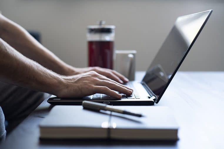 Mains sur un clavier d’ordinateur près d’un carnet et d’une tasse, poste de travail minimaliste pour faire du télétravail à domicile en journée.