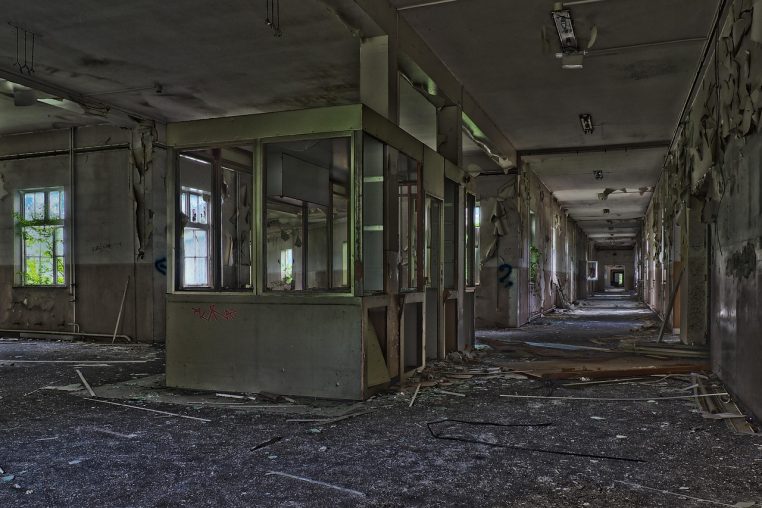 Couloir intérieur abandonné avec portes ouvertes et lumière naturelle au fond, dans un ancien bâtiment hospitalier.