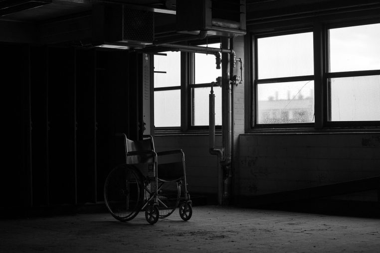 Fauteuil roulant isolé dans une pièce sombre d’un ancien hôpital abandonné, avec de grandes fenêtres poussiéreuses en arrière-plan.