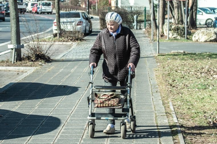 Femme âgée avançant seule avec un déambulateur sur un trottoir urbain, illustrant les défis du vieillissement et de la perte d’autonomie.