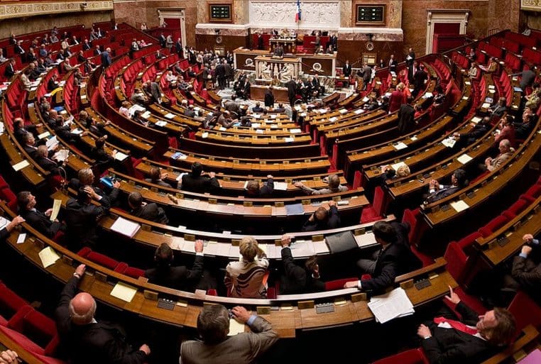 Vue large de l’hémicycle de l’Assemblée nationale rempli de députés, pendant une séance de travail sur les bancs rouges.