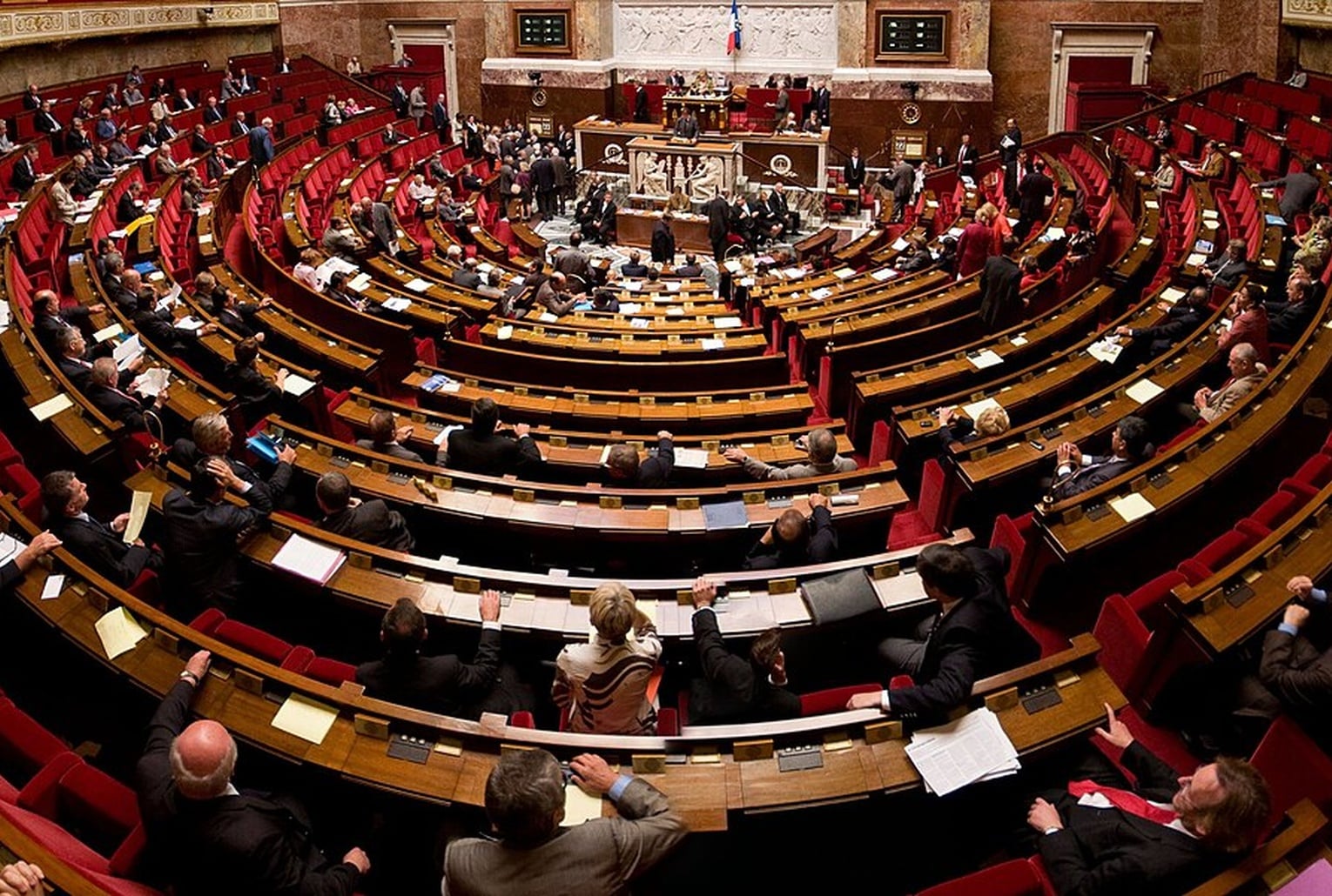 Vue large de l’hémicycle de l’Assemblée nationale rempli de députés, pendant une séance de travail sur les bancs rouges.