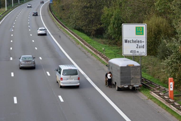 Utilitaire gris immobilisé sur la bande d’arrêt d’urgence d’une autoroute à plusieurs voies, sous un grand panneau de signalisation indiquant une sortie à venir